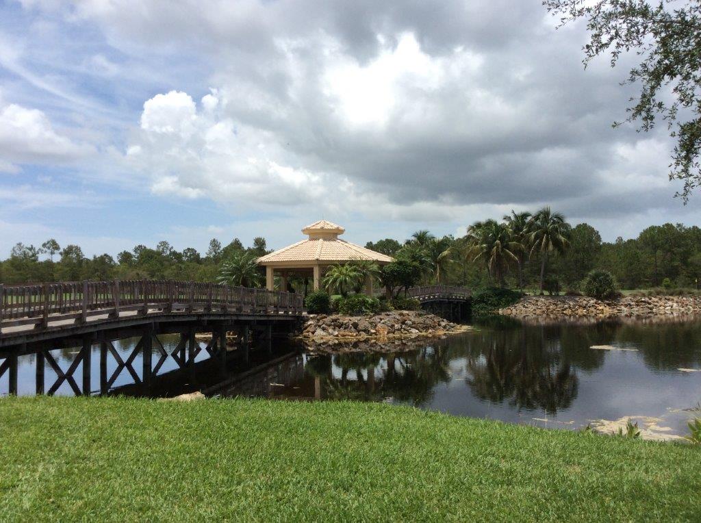 Wooden bridge over a calm pond with a gazebo and lush greenery under a cloudy sky.