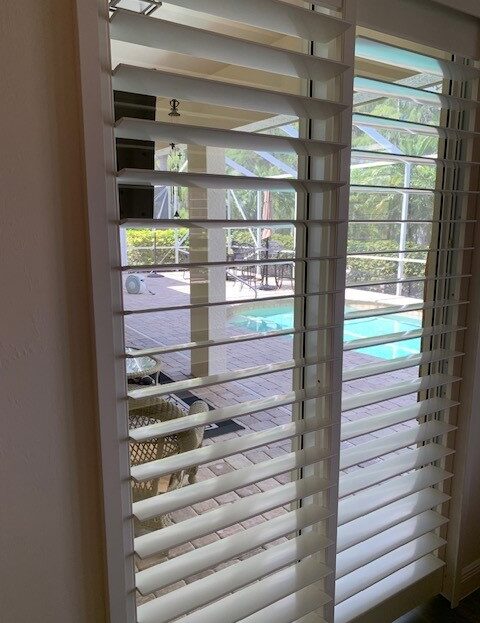 View through white blinds showing a backyard pool and patio area.