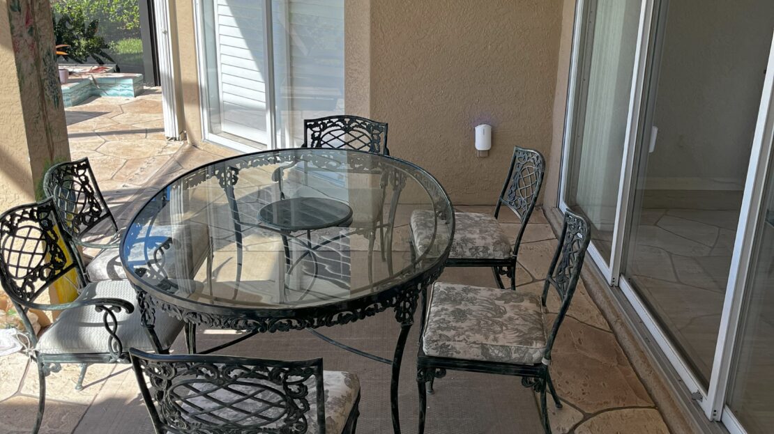 Outdoor patio with glass table and four metal chairs in a sunny backyard.