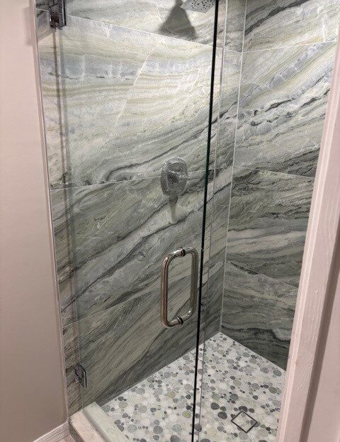 Modern glass shower door with marble walls and pebble floor.