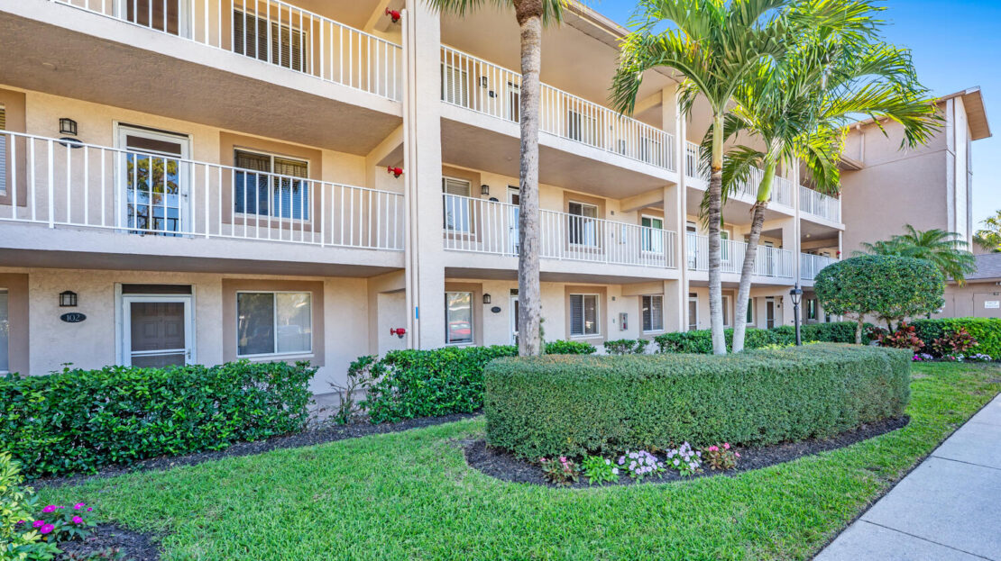Sunny exterior view of a beige multi-unit apartment building with balconies and palm trees.