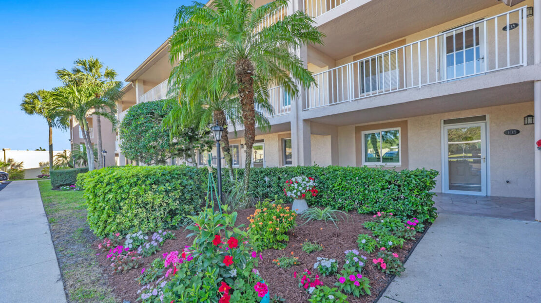 Colorful flower garden in front of a beige apartment building with palm trees.