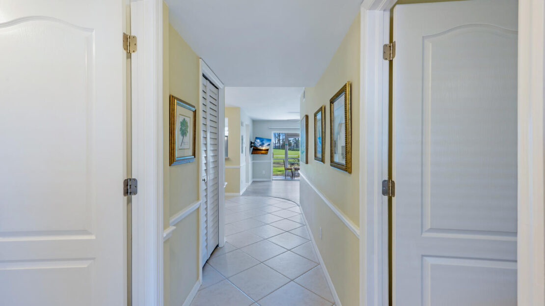 Bright hallway with framed artwork and white doors leading to a sunlit living area.