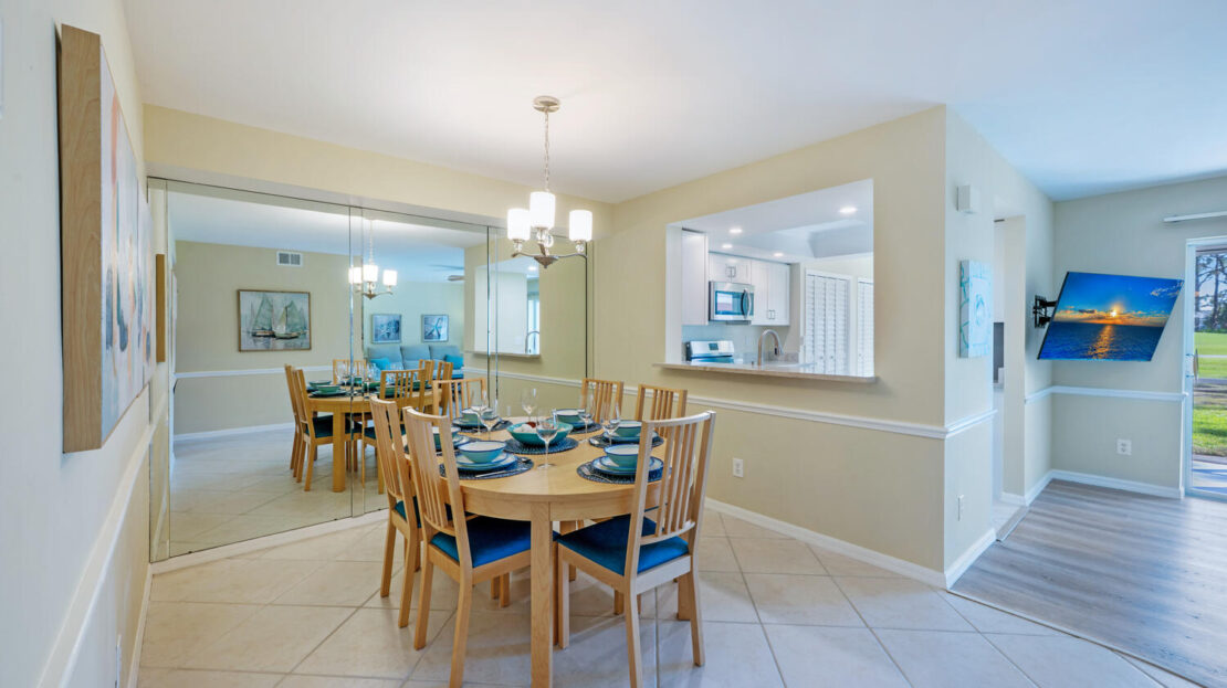 Bright dining room with wooden table set for six and a view into the kitchen.