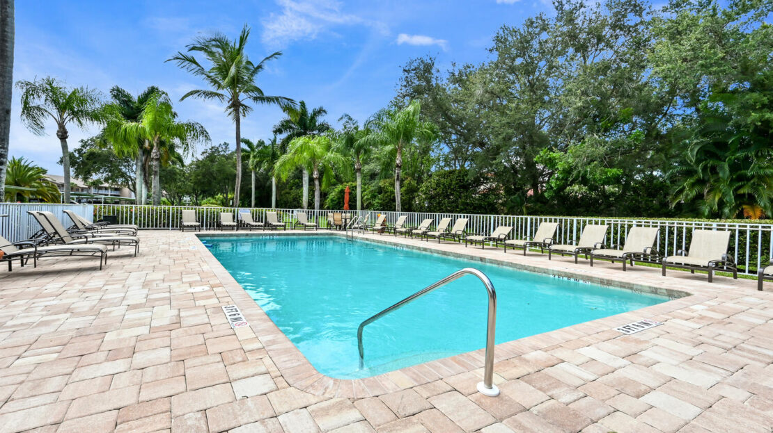 Outdoor swimming pool surrounded by lounge chairs and palm trees under a clear blue sky.