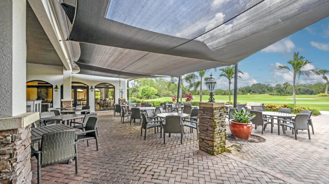 Outdoor dining area with tables, chairs, and shade sails under a partly cloudy sky.