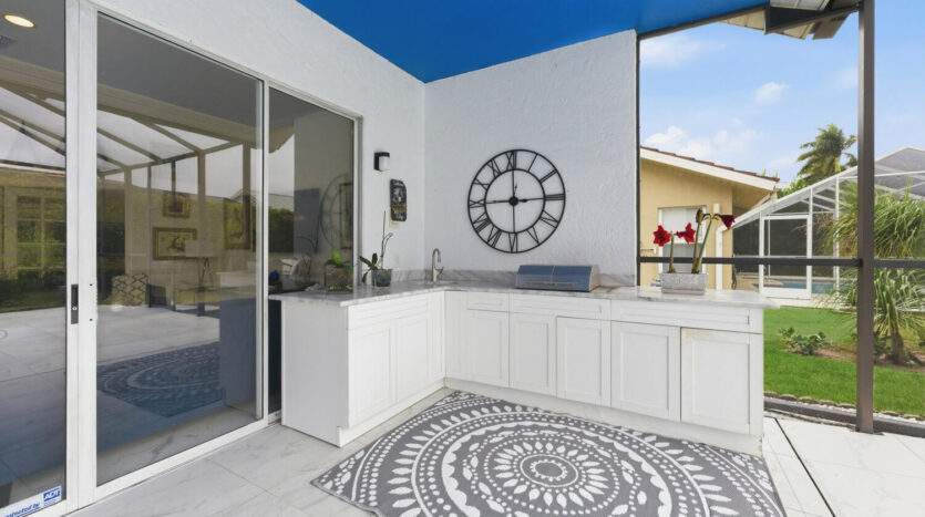 Modern outdoor kitchen with white cabinetry and a decorative wall clock under a clear blue sky.