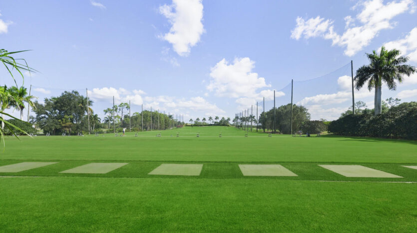 A lush green cricket field under a bright blue sky with scattered clouds.